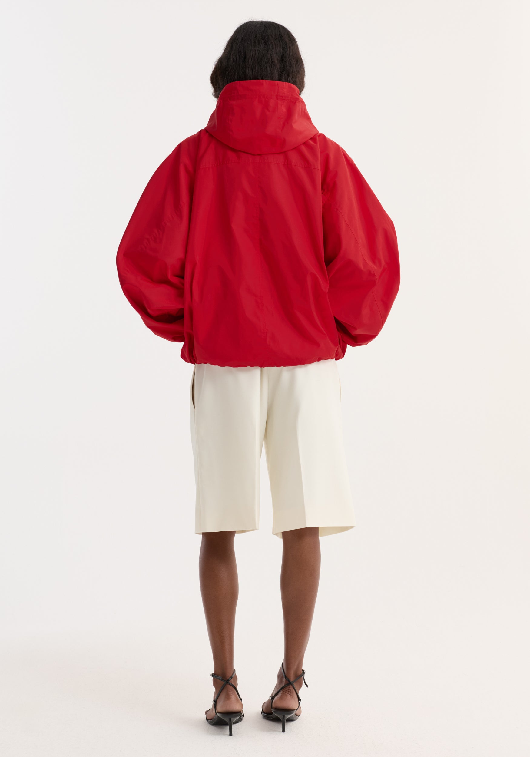 Rear view of a person wearing a bright red Volume silk-blend anorak | red with a high collar and voluminous sleeves paired with off-white shorts and black strappy heels against a plain white background.