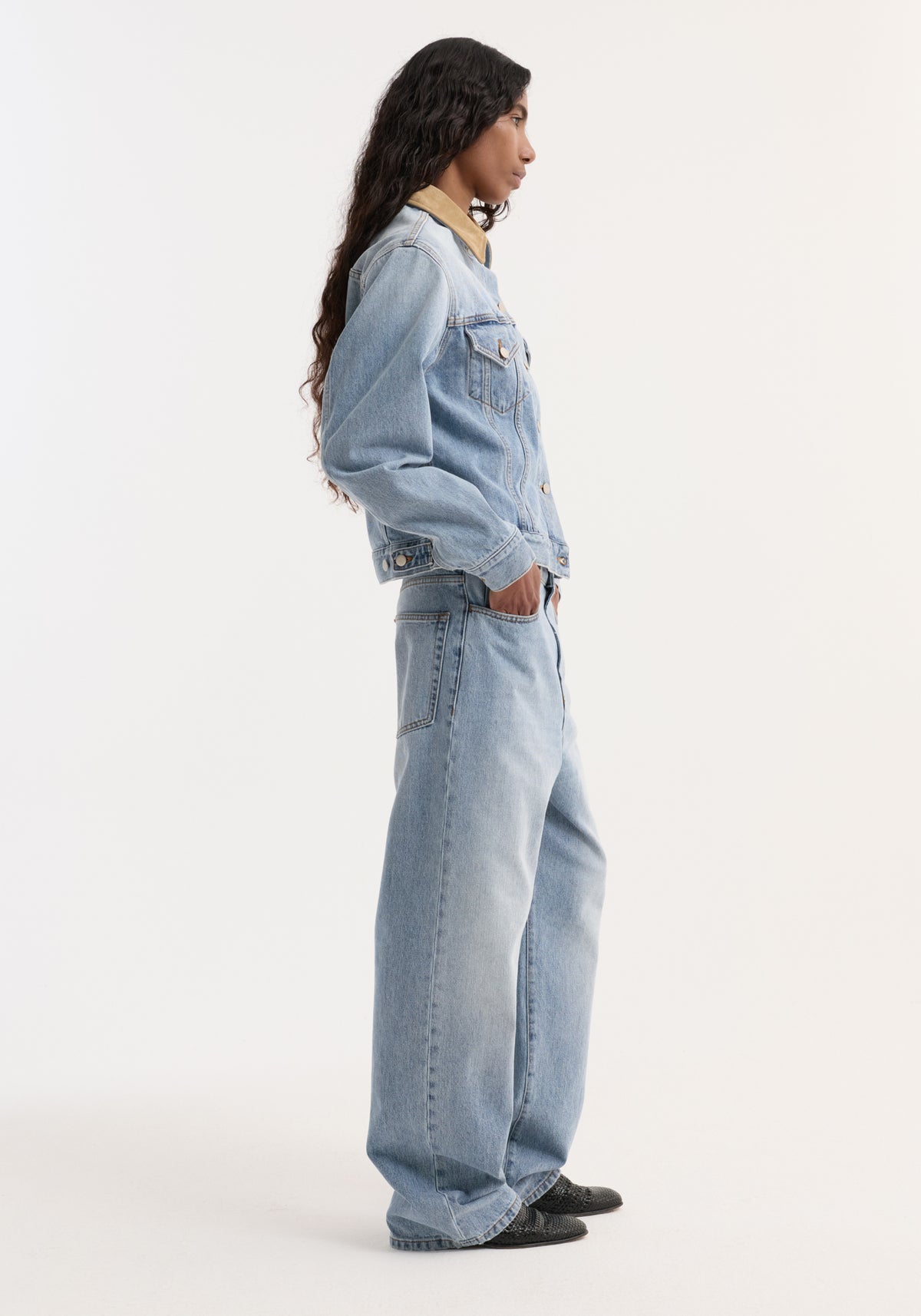 Woman wearing a Suede collar fitted denim jacket | classic blue, standing in profile with hands in wide-leg denim jeans pockets against a plain white background.