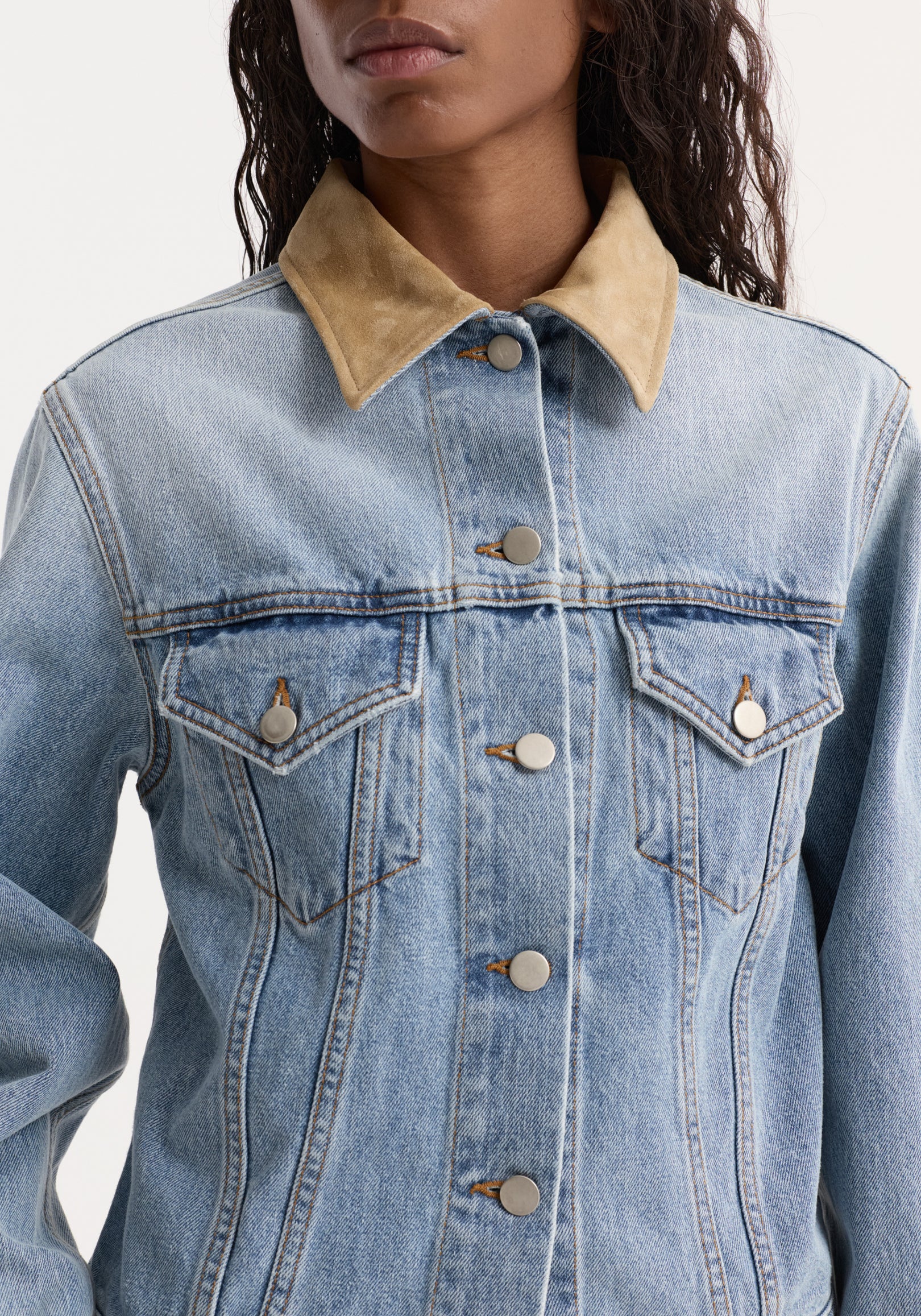 Close-up of a fitted classic blue denim jacket with a contrasting tan suede collar, button closures, and chest flap pockets featuring metal buttons, worn by a model.