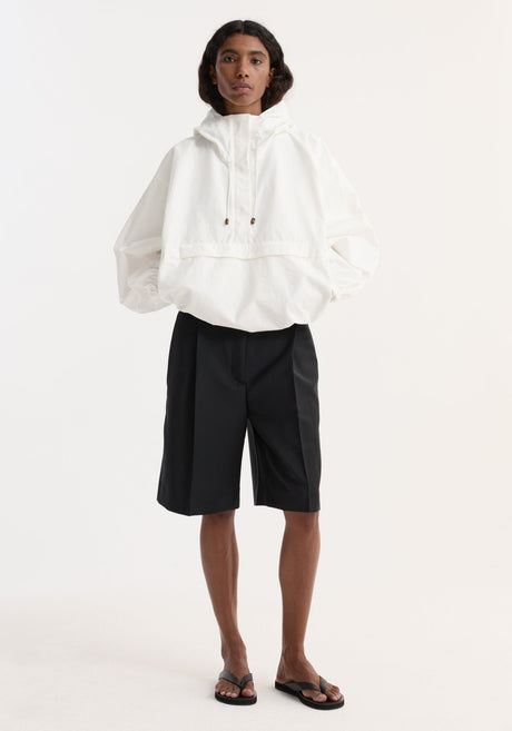 Model wearing an oversized optic white textured cotton-blend anorak with hood, front and side pockets, paired with black knee-length shorts and flip-flop sandals.