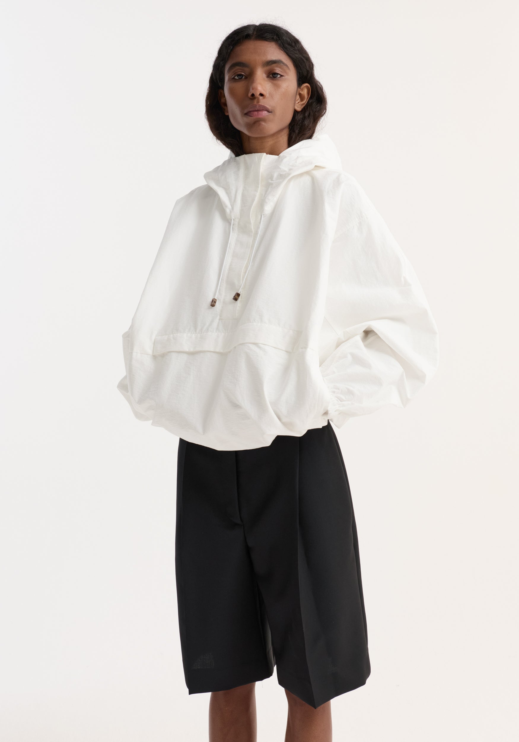 Woman wearing a loose fit textured cotton-blend anorak | optic white with a high collar and drawstring hood, paired with black knee-length shorts against a white background.
