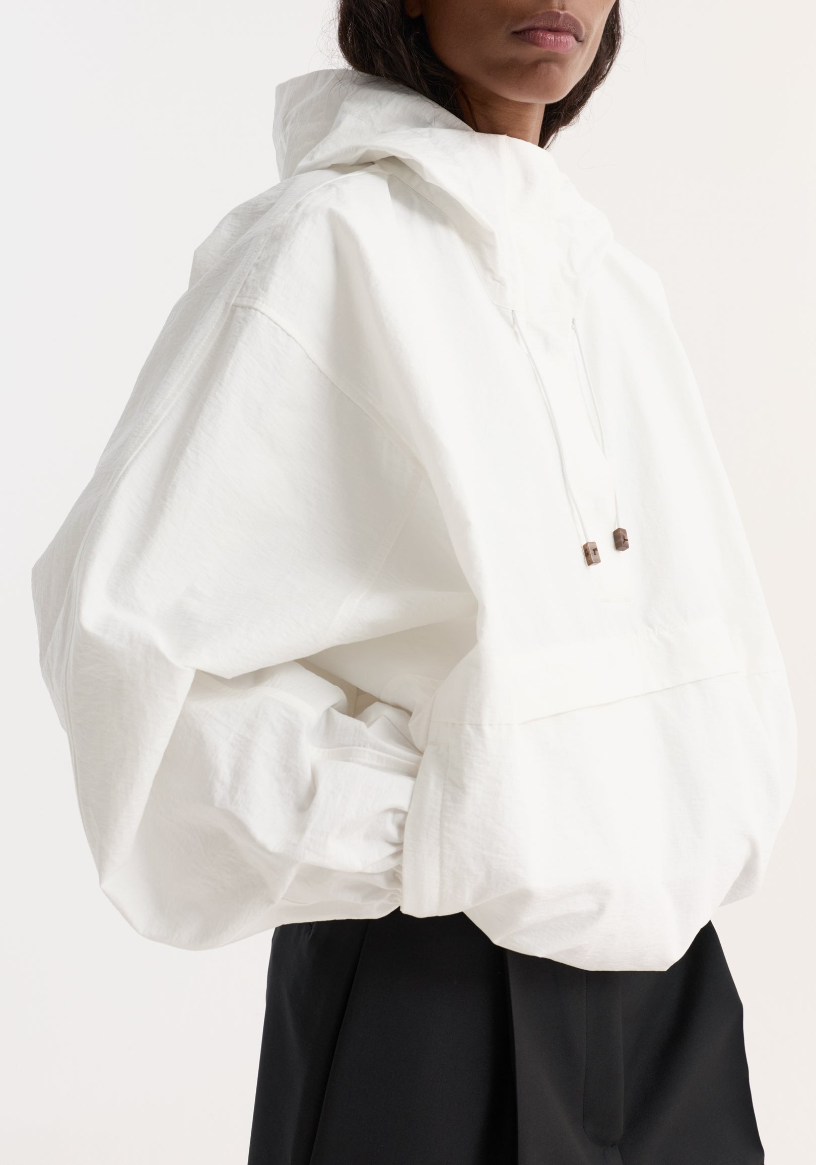 Close-up of a model wearing a textured cotton-blend anorak | optic white with a hood, loose fit, and long sleeves, paired with black pants.