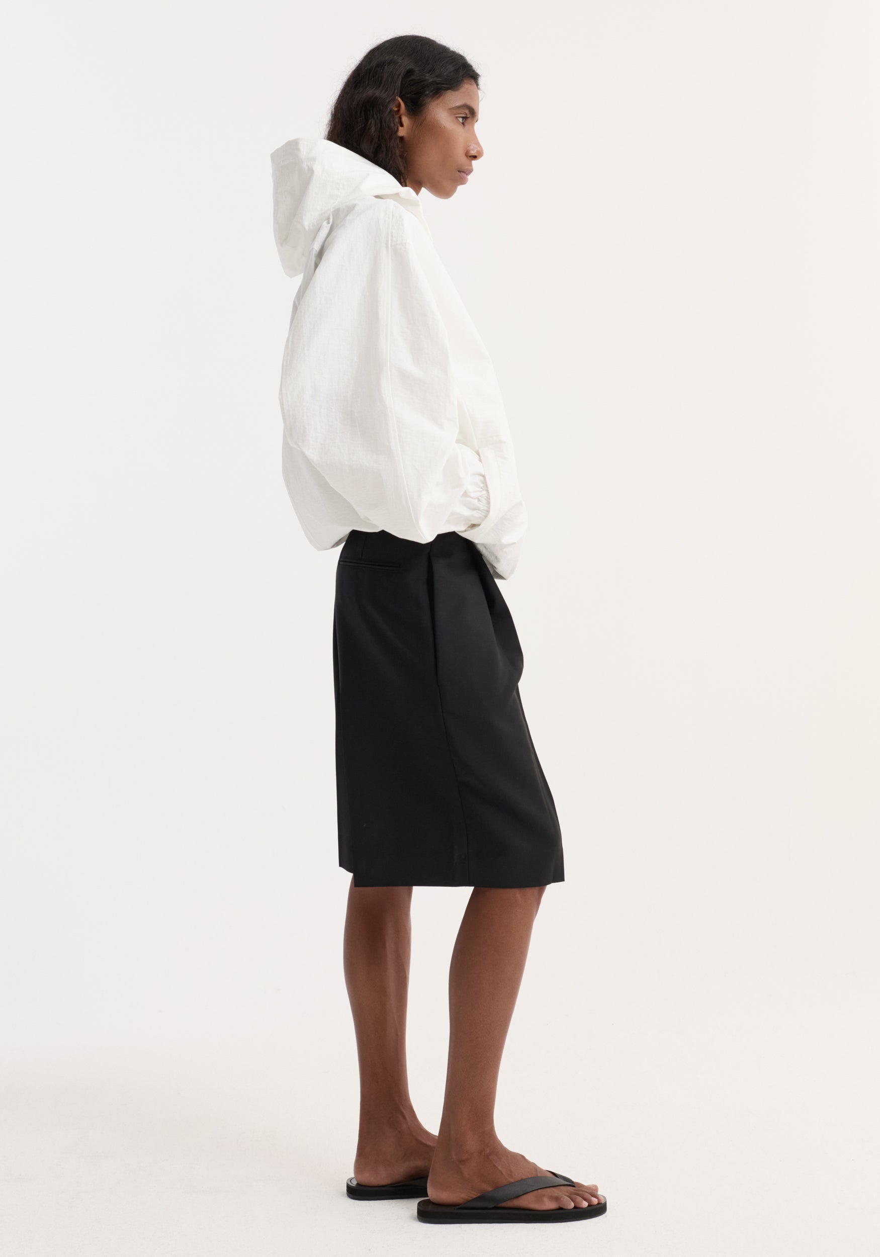 Side view of a woman wearing a textured cotton-blend anorak | optic white with a hood, paired with a black knee-length skirt and black sandals against a plain background.