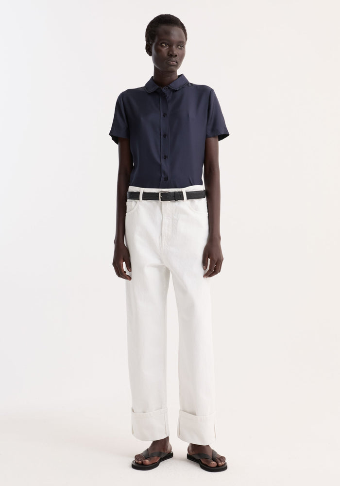 Person wearing Embroidered silk fitted shirt with short sleeves and embroidery, tucked into high-waisted trousers, accessorized with a black belt and sandals.