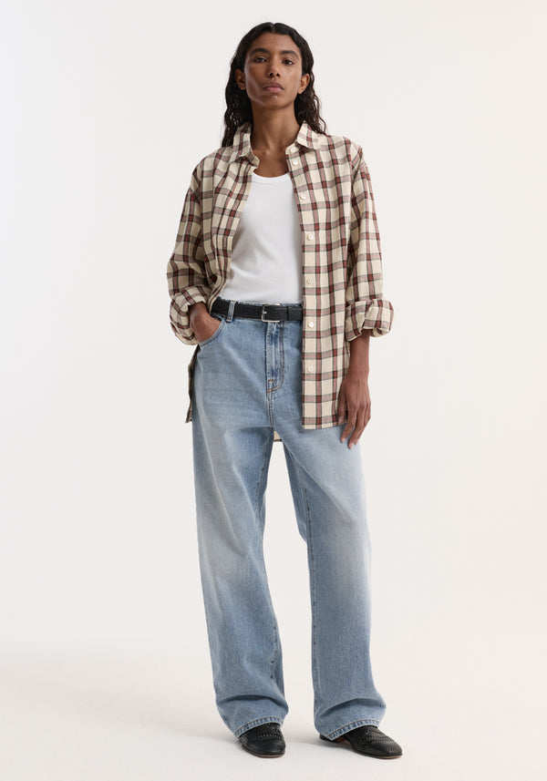 Model wearing an oversized check shirt, red table check pattern, styled open over a white tank, paired with light-wash jeans and black slip-on shoes.