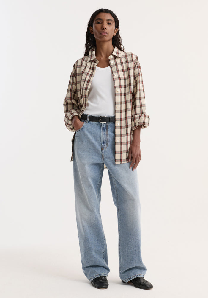 Model wearing an oversized check shirt, red table check pattern, styled open over a white tank, paired with light-wash jeans and black slip-on shoes.