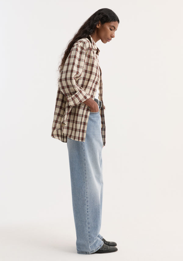 Side-profile of a person wearing an oversized check shirt in a retro pattern, styled untucked with light-wash jeans and black woven slip-on shoes.