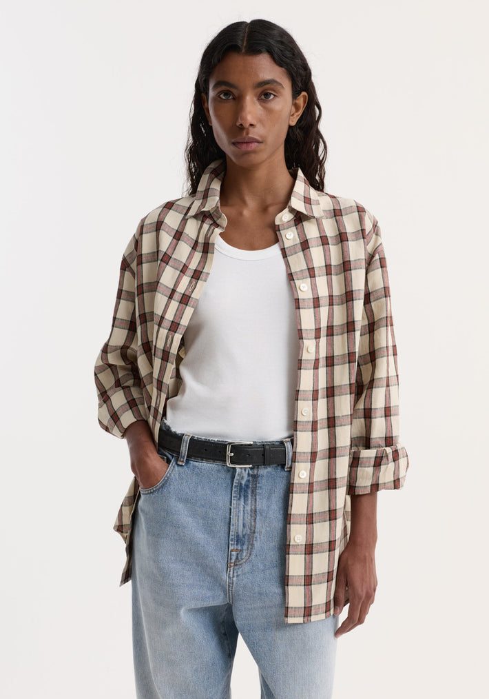 Model in an oversized check shirt, red table check pattern, unbuttoned over a white tank, paired with light-wash jeans and a black belt.