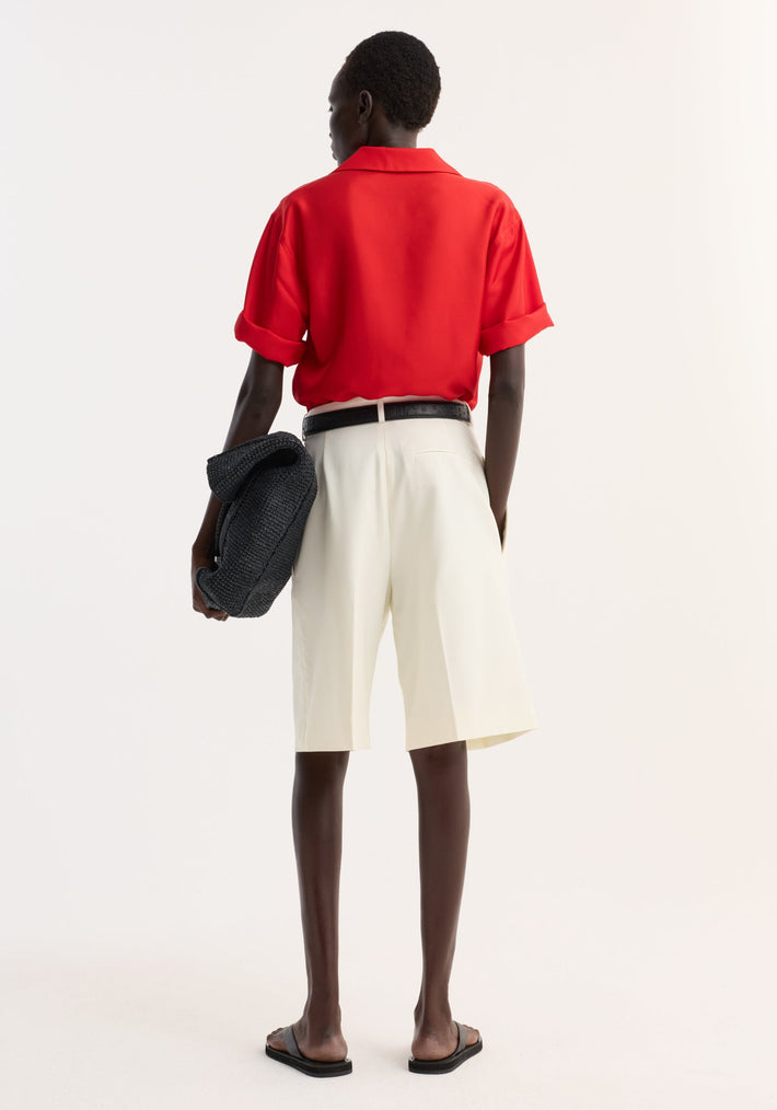 Model wearing a Camp collar silk shirt | red with rolled sleeves, paired with high-waisted ivory shorts, black belt, slide sandals, and holding a chunky-knit black clutch.