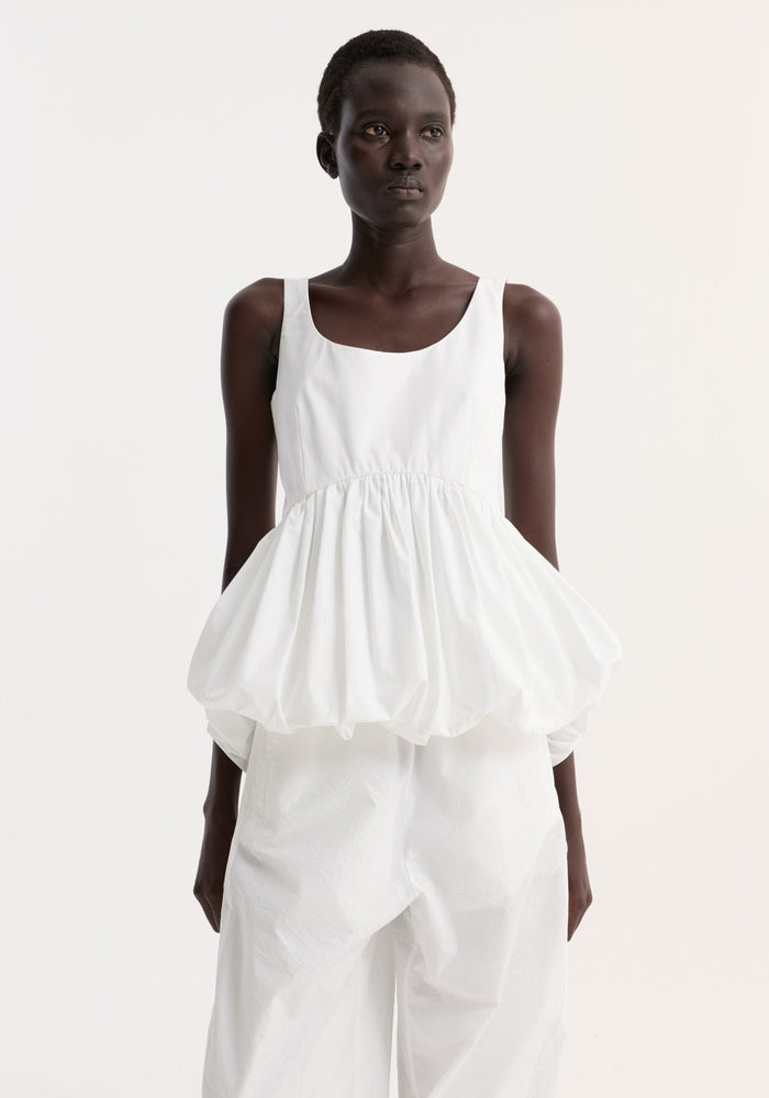 Voluminous peplum top in optic white, featuring a sleeveless scoop neck and low back, worn by a model with loose, wide-leg trousers.