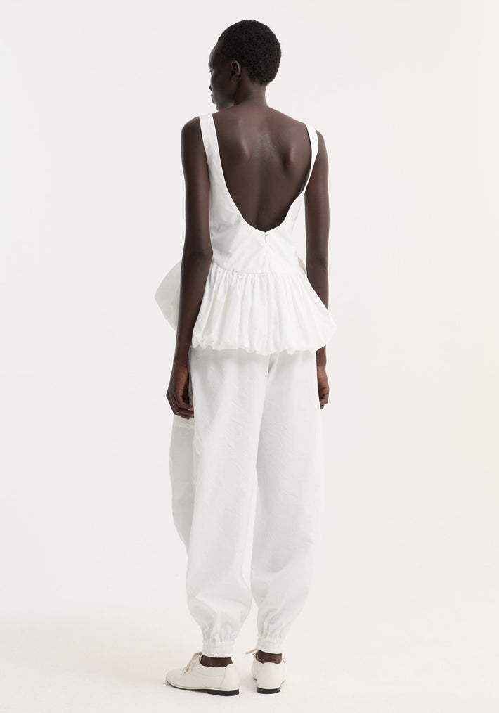 Model wearing Voluminous peplum top | optic white, featuring a sleeveless, low-back design with a gathered peplum hem, paired with loose-fitting trousers and white sneakers.