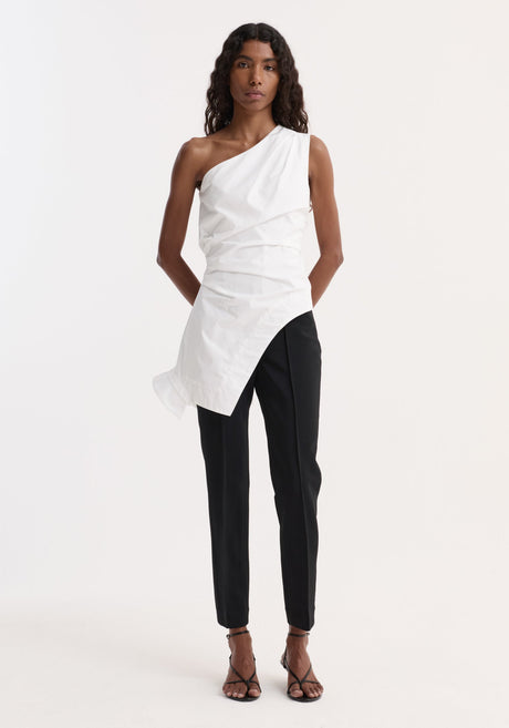 Model in Reimagined one shoulder top, optic white, with asymmetric drape and inverted collar, paired with tailored black trousers and strappy sandals, hands behind back.