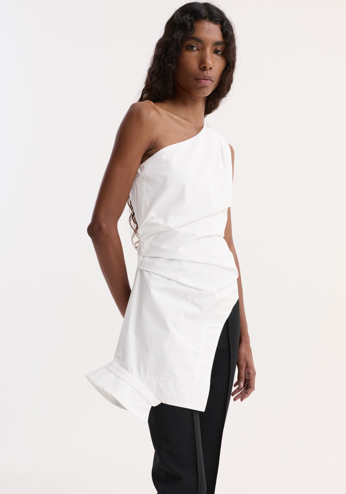 Model wears Reimagined one shoulder top in optic white, showcasing an asymmetrical drape, fitted waist, and deconstructed collar, paired with straight-leg black pants.