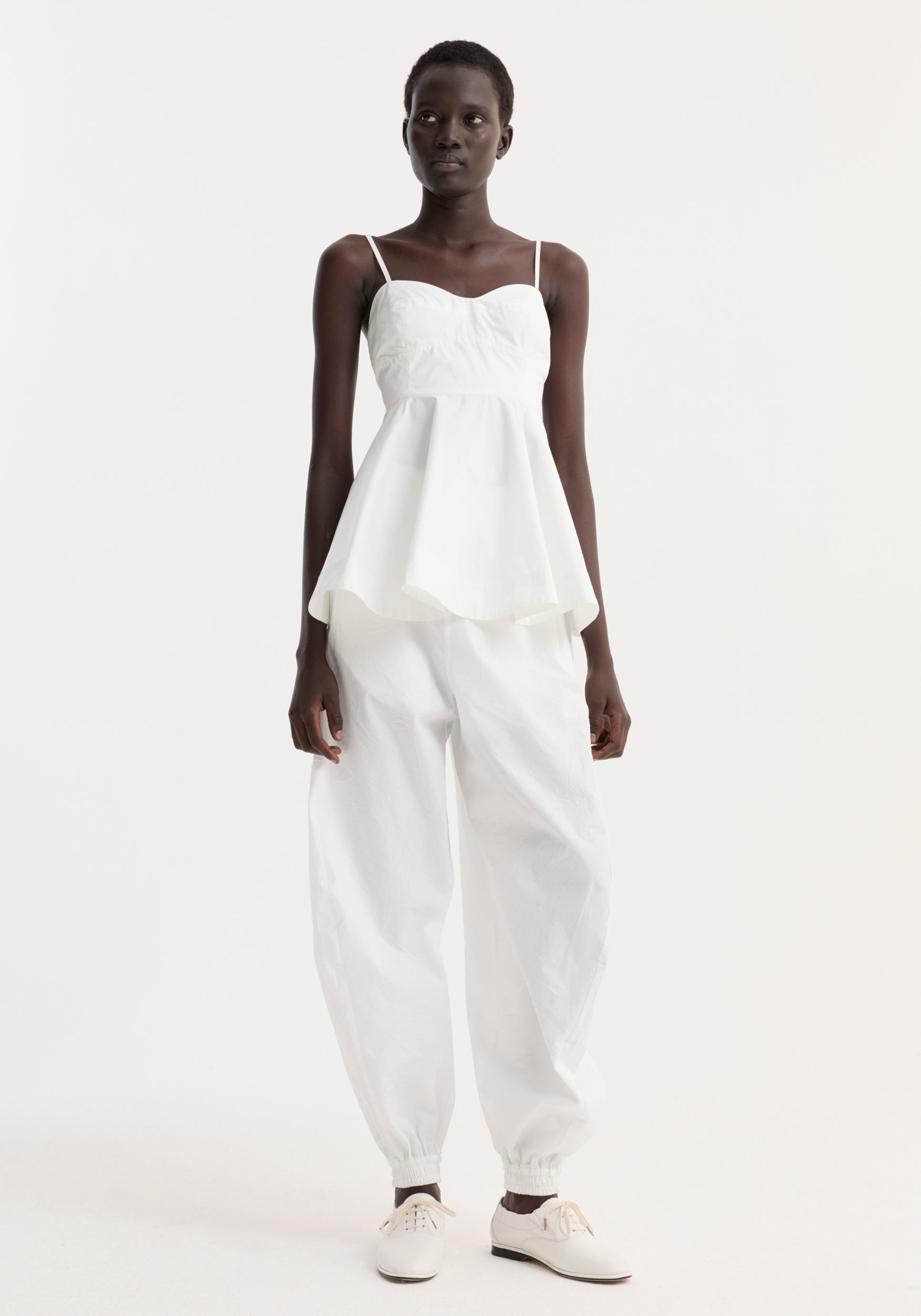 A model wearing a bustier peplum top | optic white, featuring thin straps and a structured, flared waist paired with loose-fitting white pants and white lace-up shoes.
