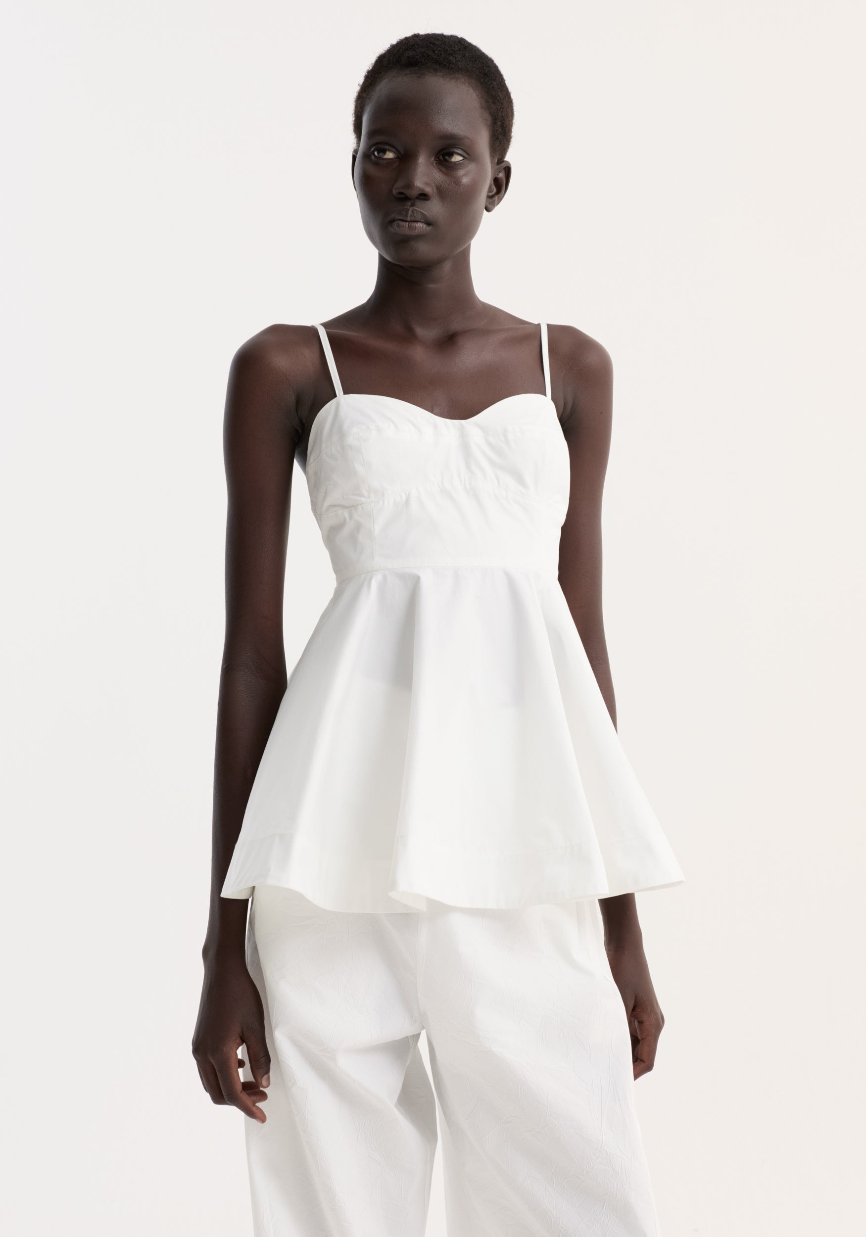 Woman wearing a Bustier peplum top | optic white with thin straps and a fitted bodice that flares at the waist, paired with loose white pants against a plain light background.
