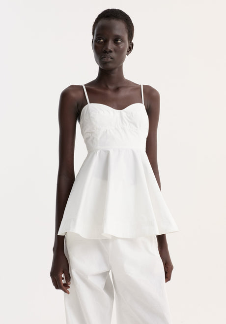Model wearing Bustier peplum top in optic white, featuring bustier detailing, flared peplum, and adjustable straps, paired with loose-fit trousers and lace-up shoes.