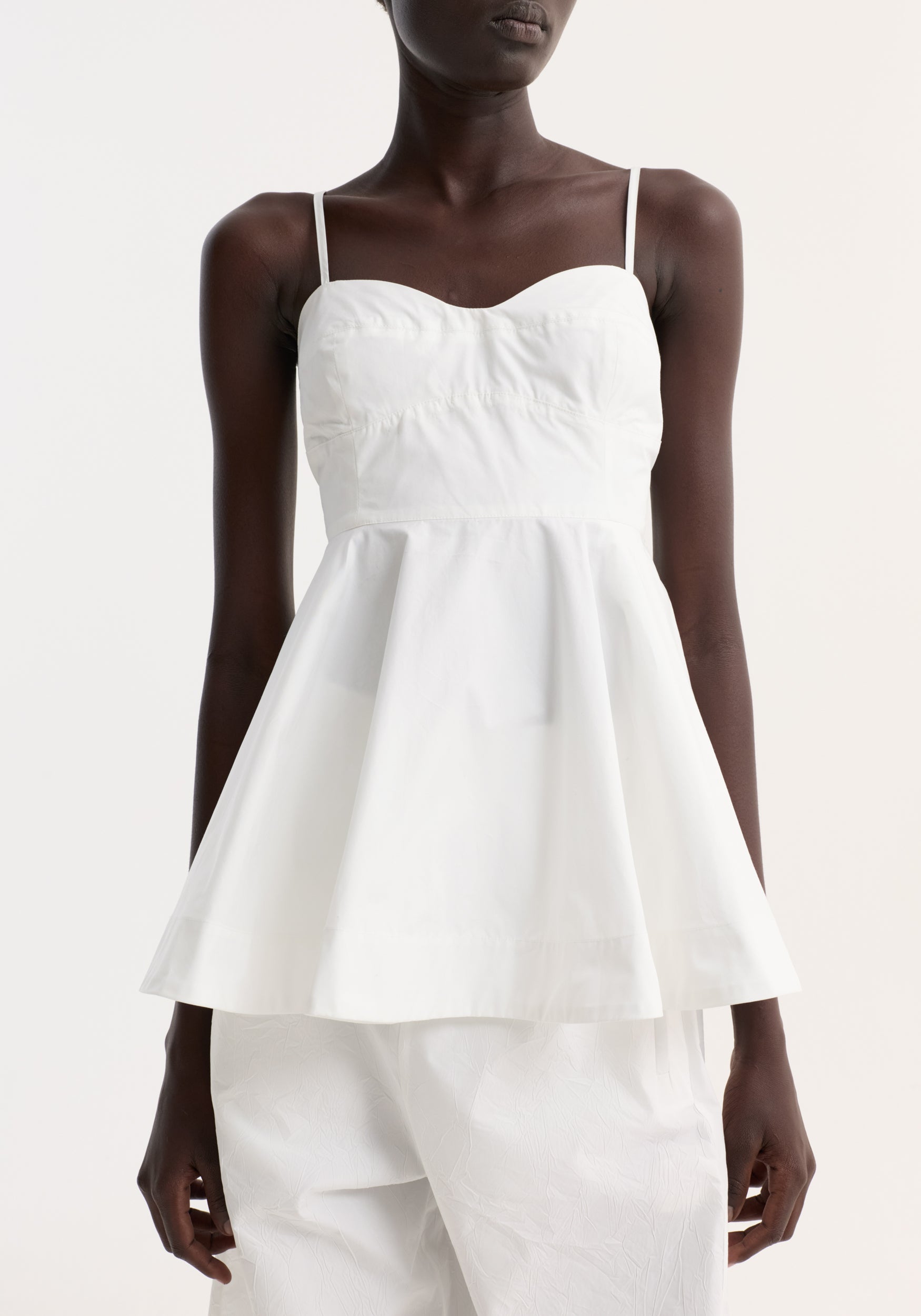 Woman wearing a white bustier peplum top | optic white with thin straps and a flared waist, paired with white pants against a plain light background.