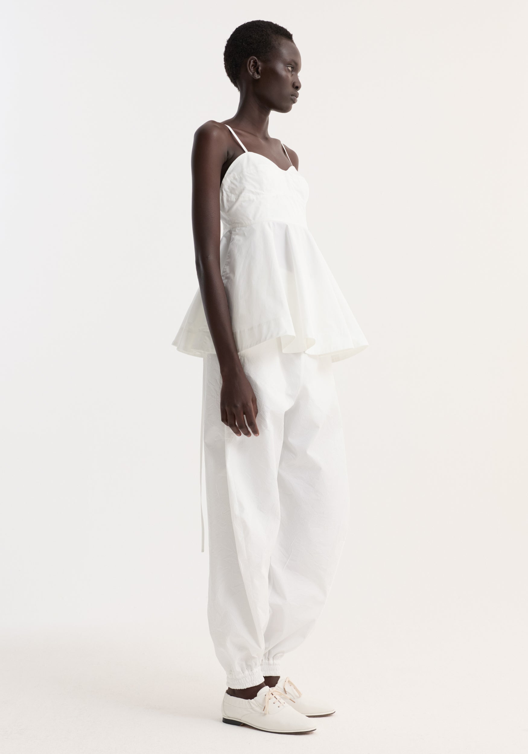 Side view of a woman wearing a white bustier peplum top | optic white with thin straps, paired with loose white pants and white shoes, standing against a plain light background.