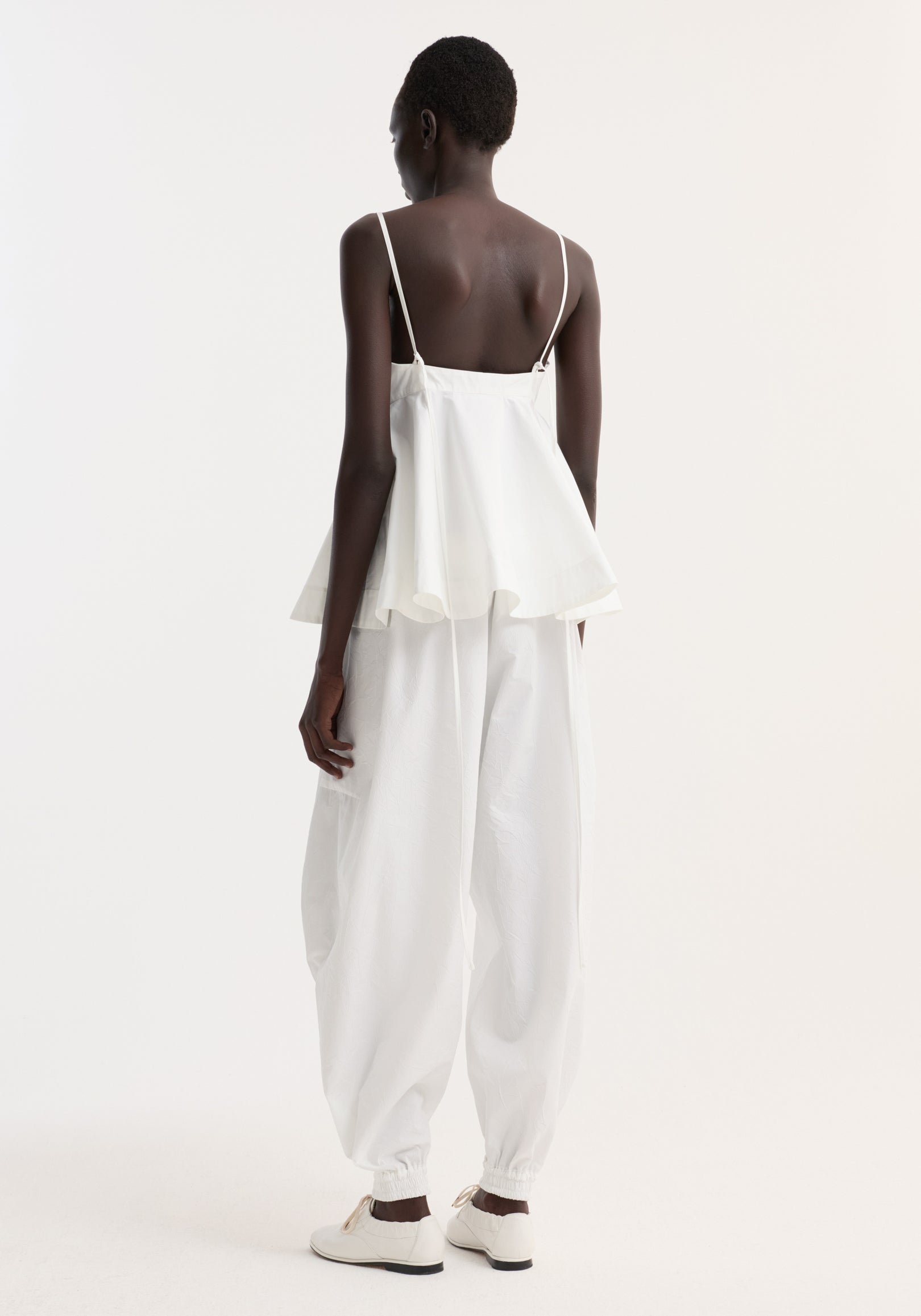 Rear view of a woman wearing a Bustier peplum top | optic white with thin straps and a flared hem, paired with loose white pants and white shoes against a plain background.