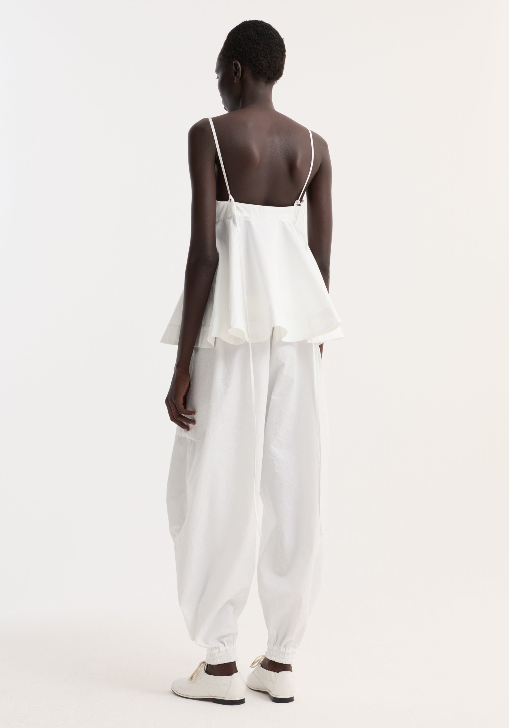 Rear view of a woman wearing a Bustier peplum top | optic white with thin straps and a flared hem, paired with loose white pants and white shoes against a plain background.