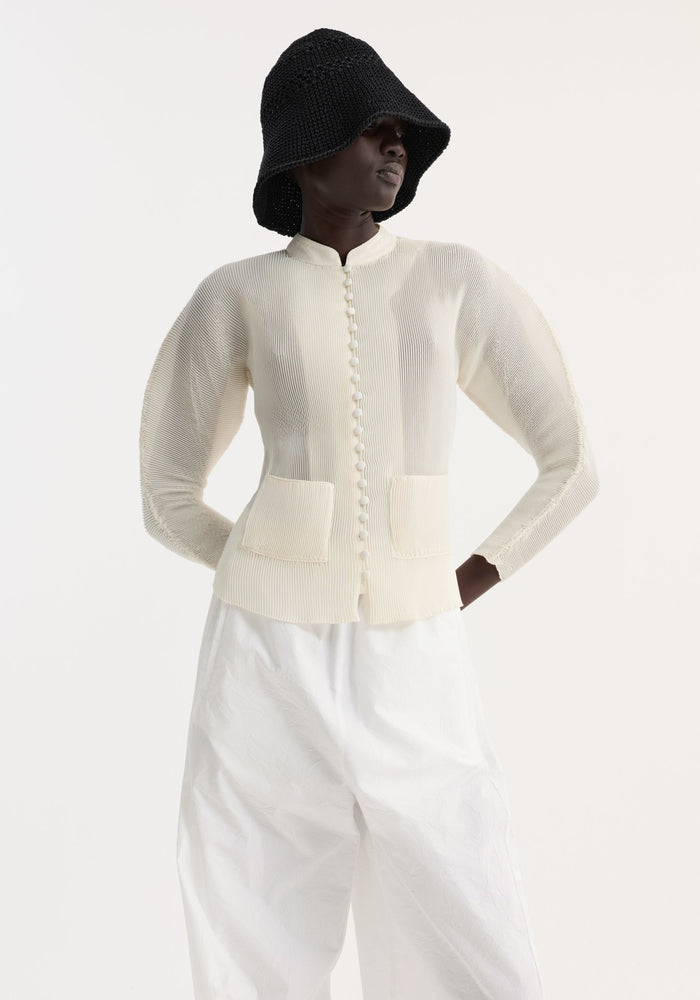 Model in cream Plissé cardigan with covered buttons, stand-up collar, cropped fit, and patch pockets, paired with white textured trousers and a black bucket hat.