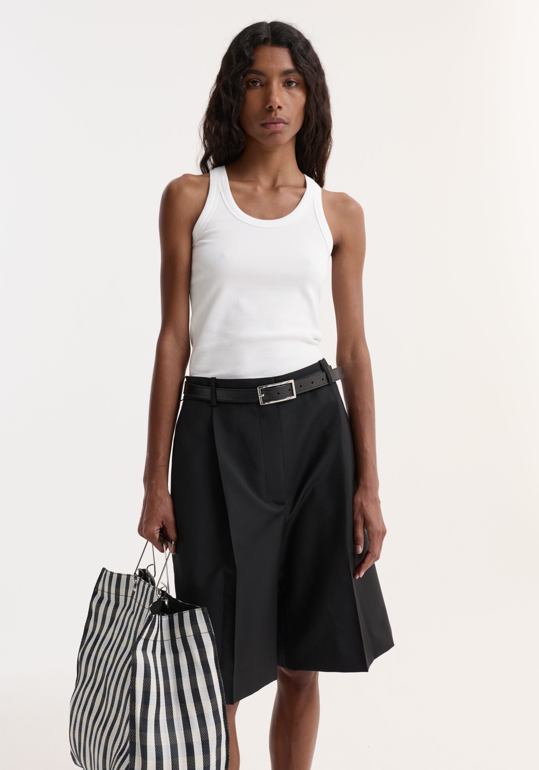 Model in fitted Scoop-neck rib top | optic white, paired with high-waisted black pleated culottes, holding a striped tote with metal ring handles.