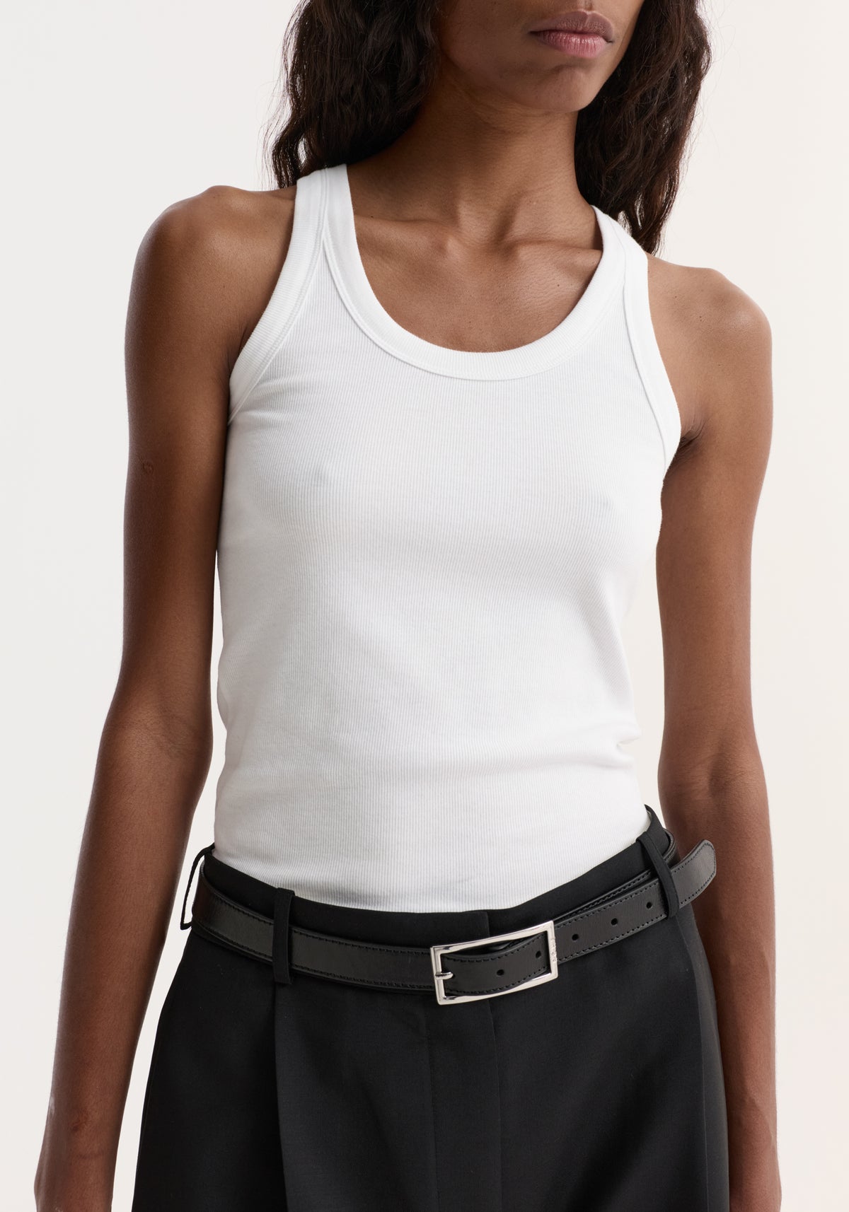 Person wearing the Scoop-neck rib top in optic white, showcasing its scoop neck and racerback design, paired with black trousers and a leather belt.