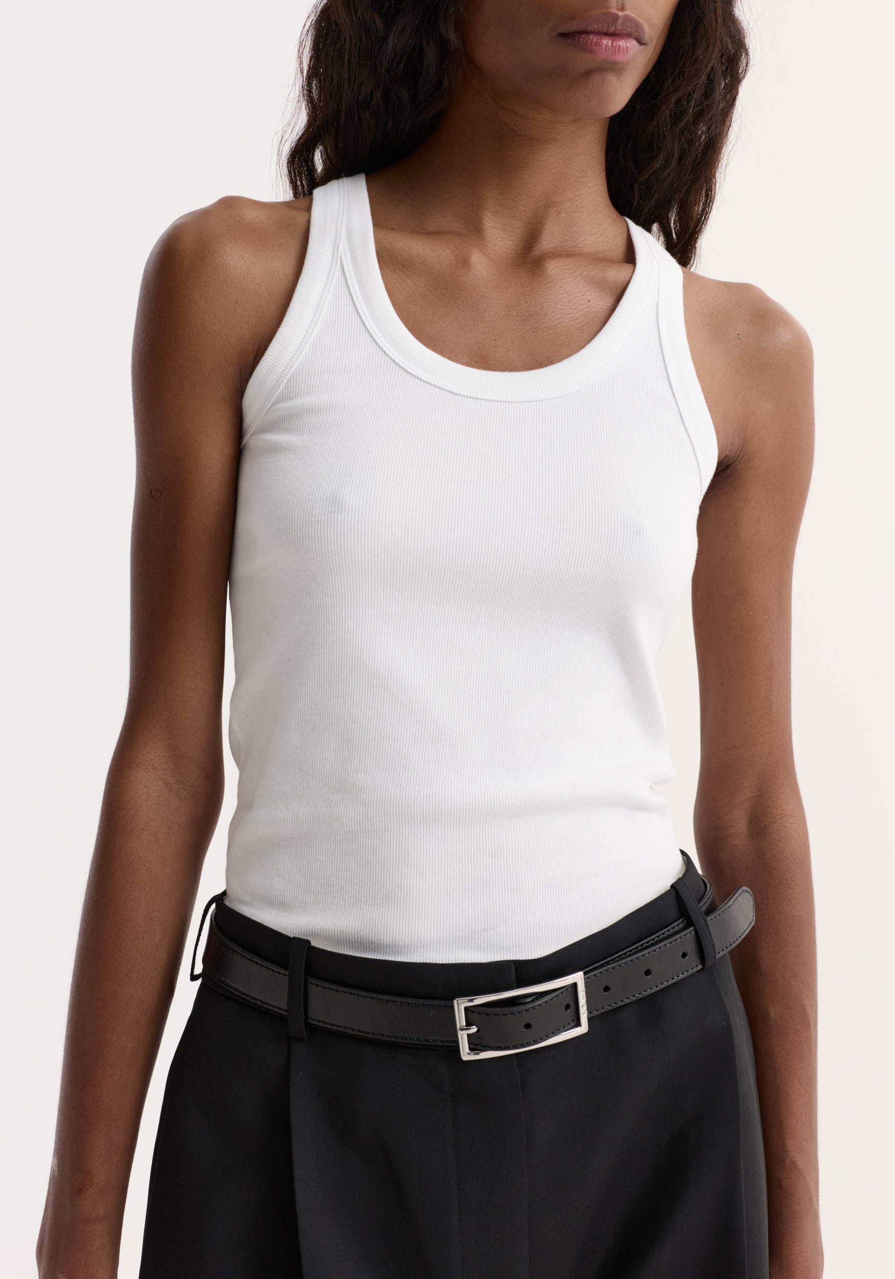 Person wearing the Scoop-neck rib top in optic white, showcasing its scoop neck and racerback design, paired with black trousers and a leather belt.
