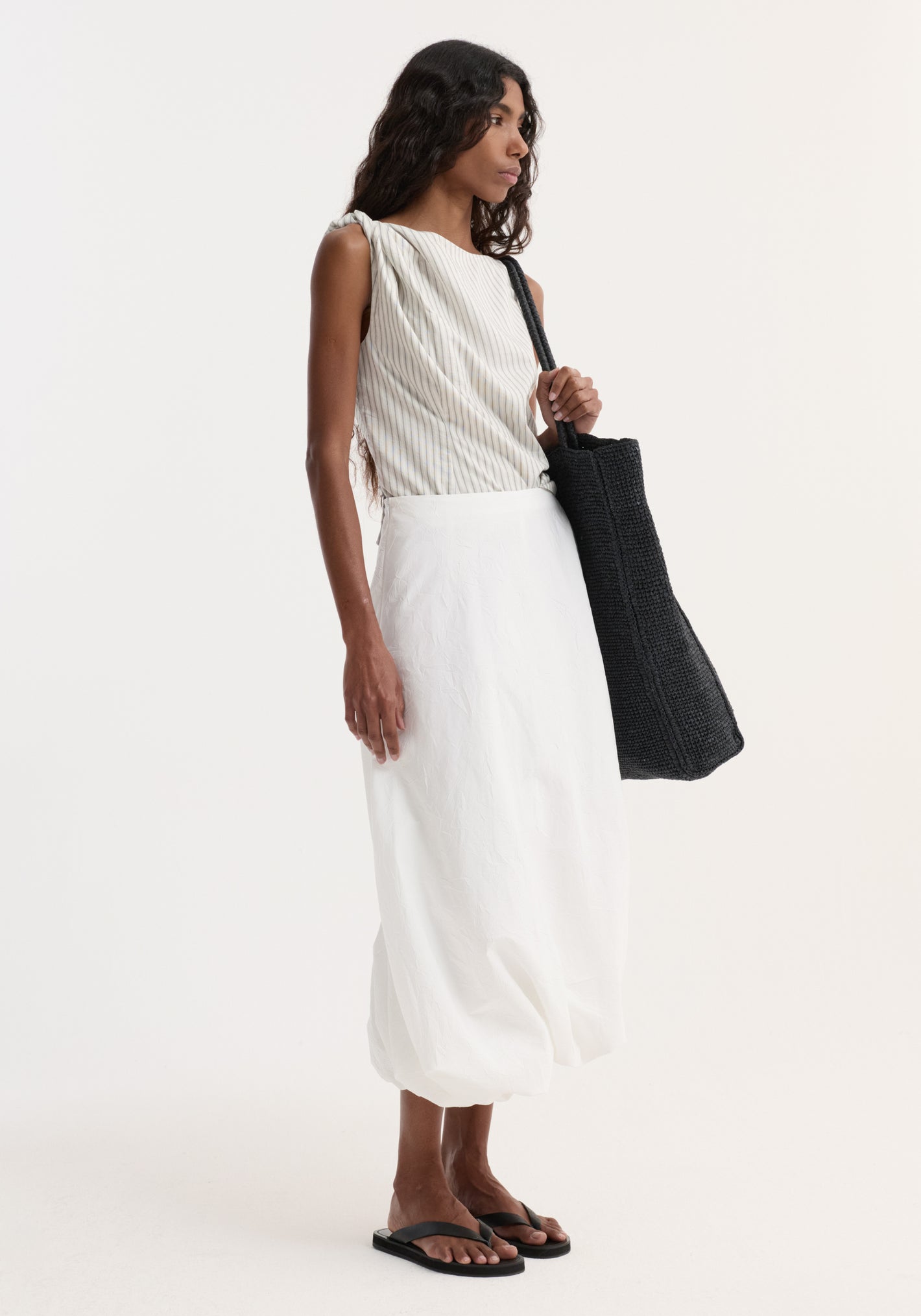 Woman wearing a Silk-blend draped top | fine stripe with twist detail on one shoulder, paired with a white balloon skirt and black sandals, holding a slouchy tote.