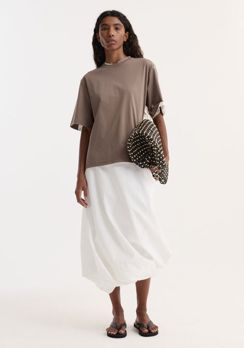 Woman wearing Oversized cotton silk t-shirt in taupe with loose fit, paired with a white voluminous skirt and black sandals, holding a woven clutch.