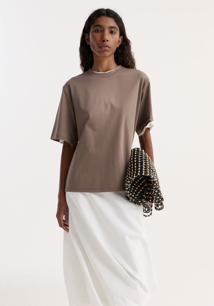 Young woman in Oversized cotton silk t-shirt | taupe, showcasing its relaxed fit and drop shoulders, paired with a woven clutch and white skirt.