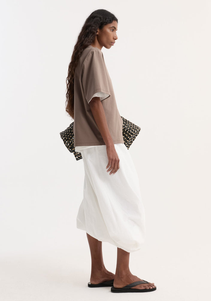Woman wearing Oversized cotton silk t-shirt in taupe with loose fit, paired with a white voluminous skirt and black sandals, holding a woven clutch.