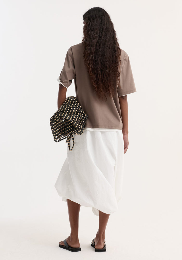 Back view of a person wearing an oversized taupe cotton-silk t-shirt with rolled sleeves, paired with a white midi skirt and black sandals.