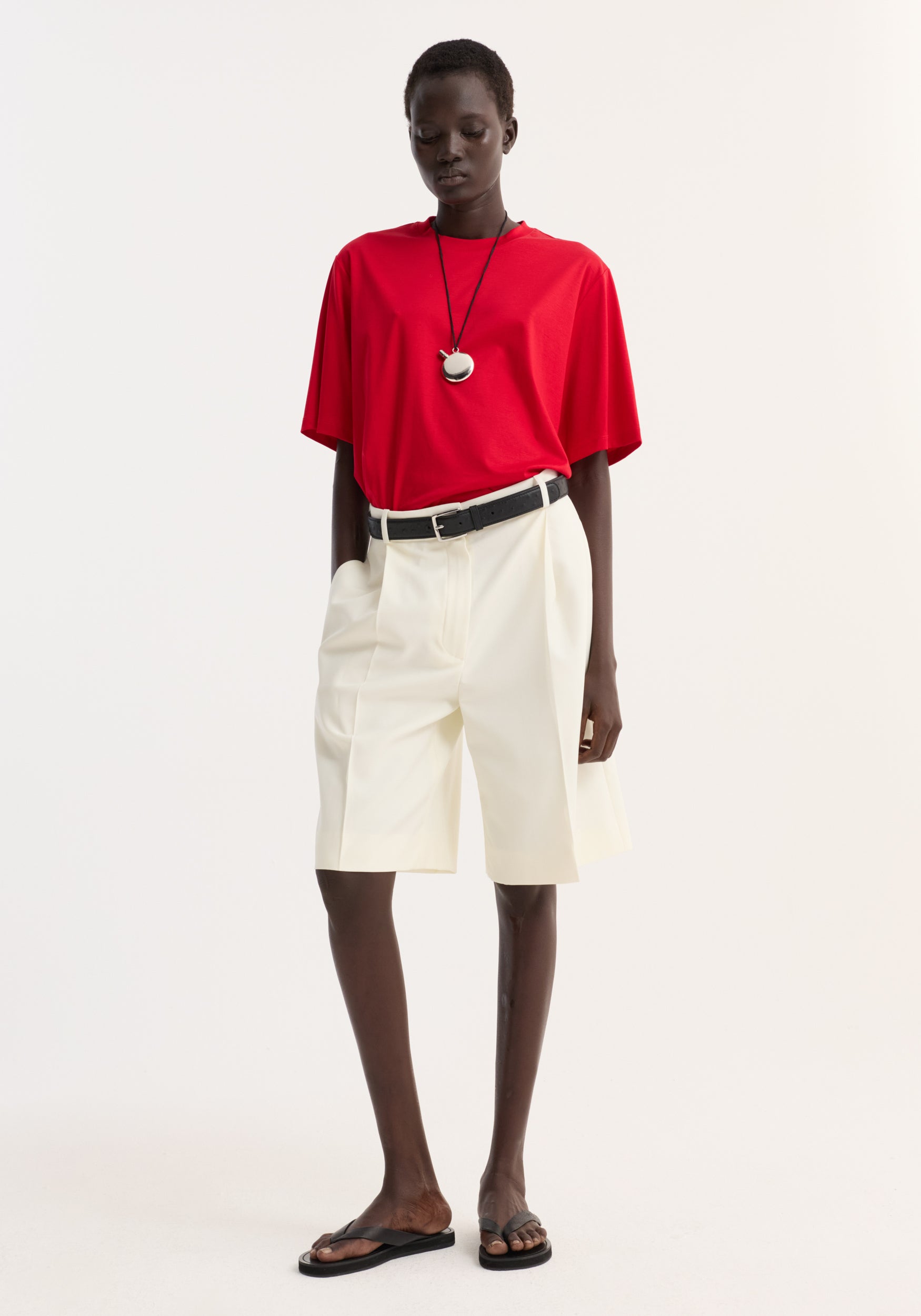 A model wearing an oversized cotton silk t-shirt | red paired with high-waisted, cream-colored shorts, a black belt, a pendant necklace, and black sandals against a plain white background.