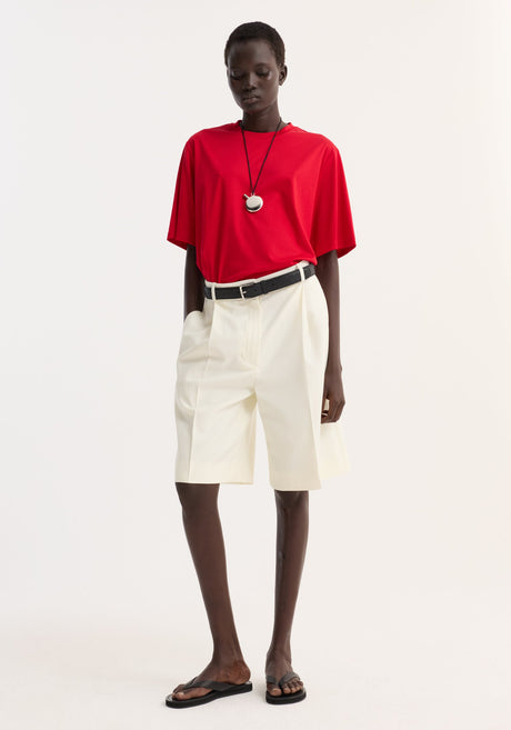 Dark-skinned model wearing an oversized cotton-silk red t-shirt with dropped shoulders, tucked into high-waisted cream pleated shorts, accessorized with a black necklace.