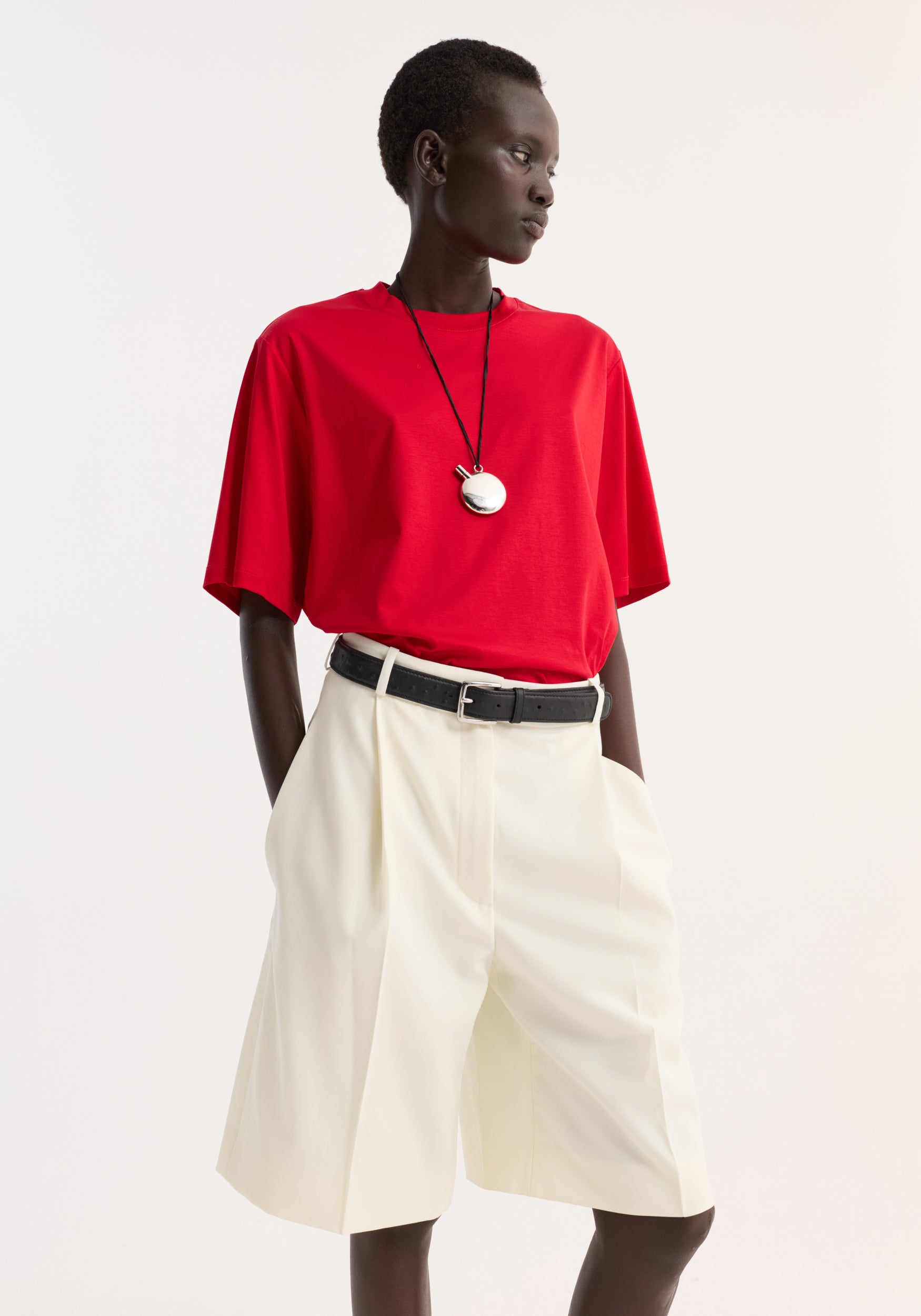 Model wearing an oversized cotton silk t-shirt | red paired with high-waisted cream shorts and a black belt, accessorized with a pendant necklace against a plain background.