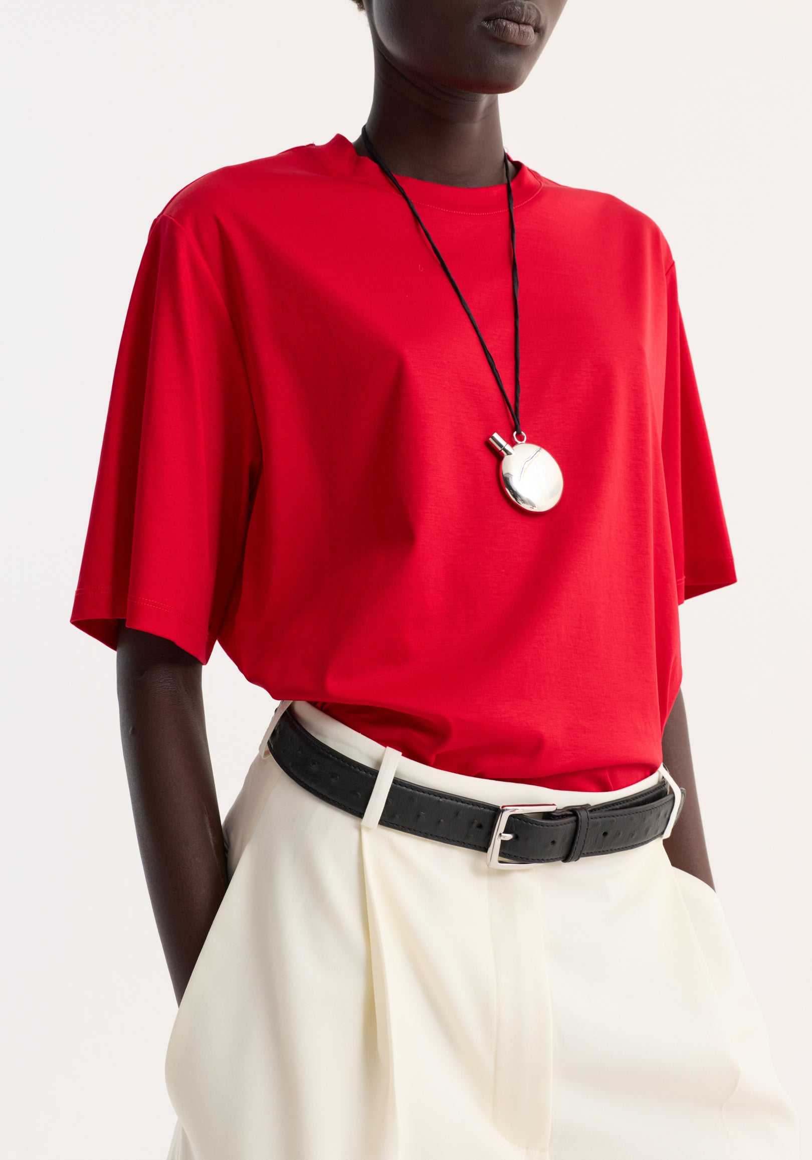 Model wearing an oversized cotton silk t-shirt | red paired with white high-waisted pants and a black belt, accessorized with a silver round pendant necklace.