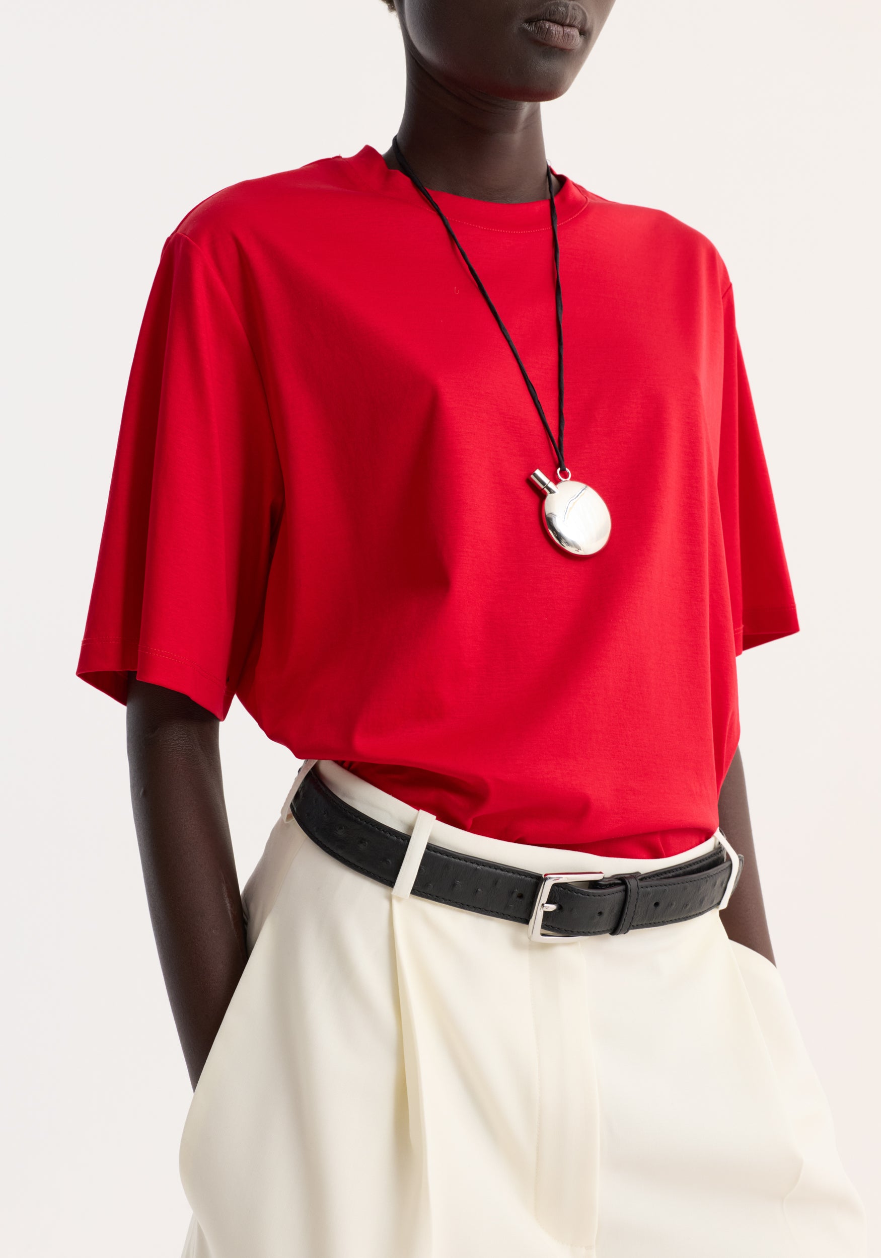 Model wearing an oversized cotton silk t-shirt | red paired with white high-waisted pants and a black belt, accessorized with a silver round pendant necklace.