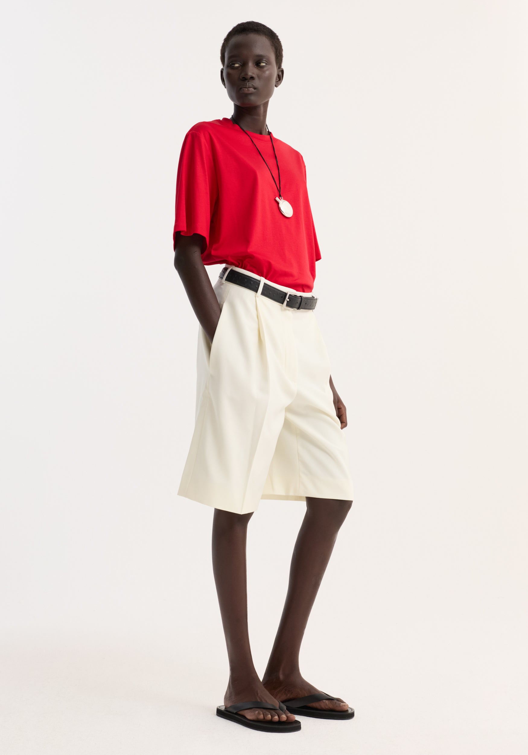 Model wearing a bright red oversized cotton silk t-shirt | red paired with cream knee-length shorts, black belt, pendant necklace, and black sandals against a plain light background.