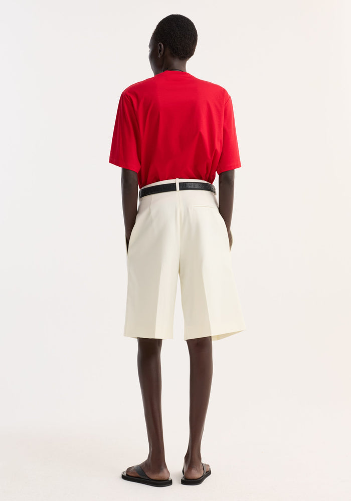 Person in an oversized red cotton-silk t-shirt with dropped shoulders and crew neck, paired with tailored off-white shorts, black belt, and slide sandals.