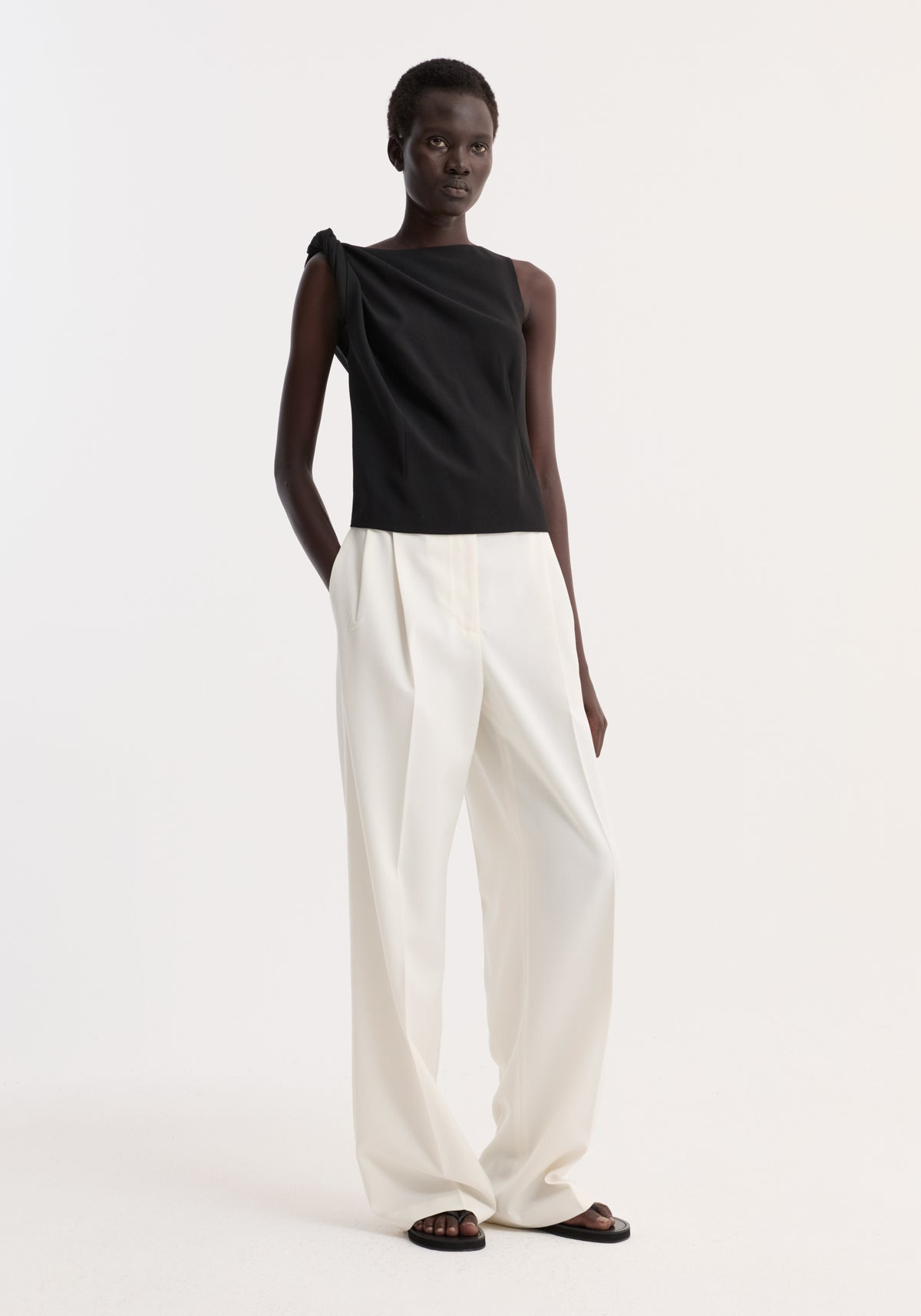 Model wearing a Draped linen silk top | black, featuring a twist detail at the shoulder, boat neck, and pleated side, paired with white trousers.