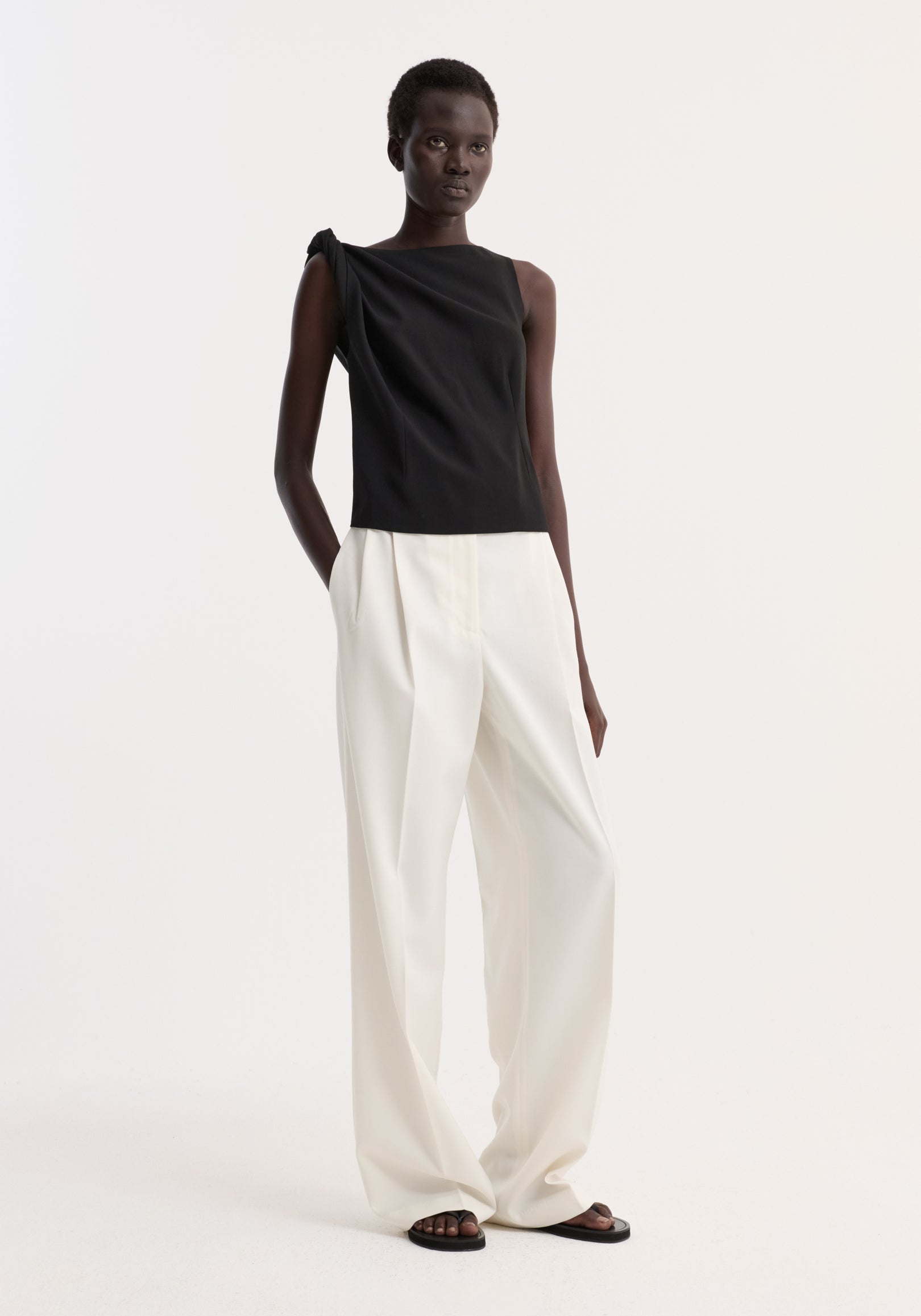 Model wearing a Draped linen silk top | black, featuring a twist detail at the shoulder, boat neck, and pleated side, paired with white trousers.