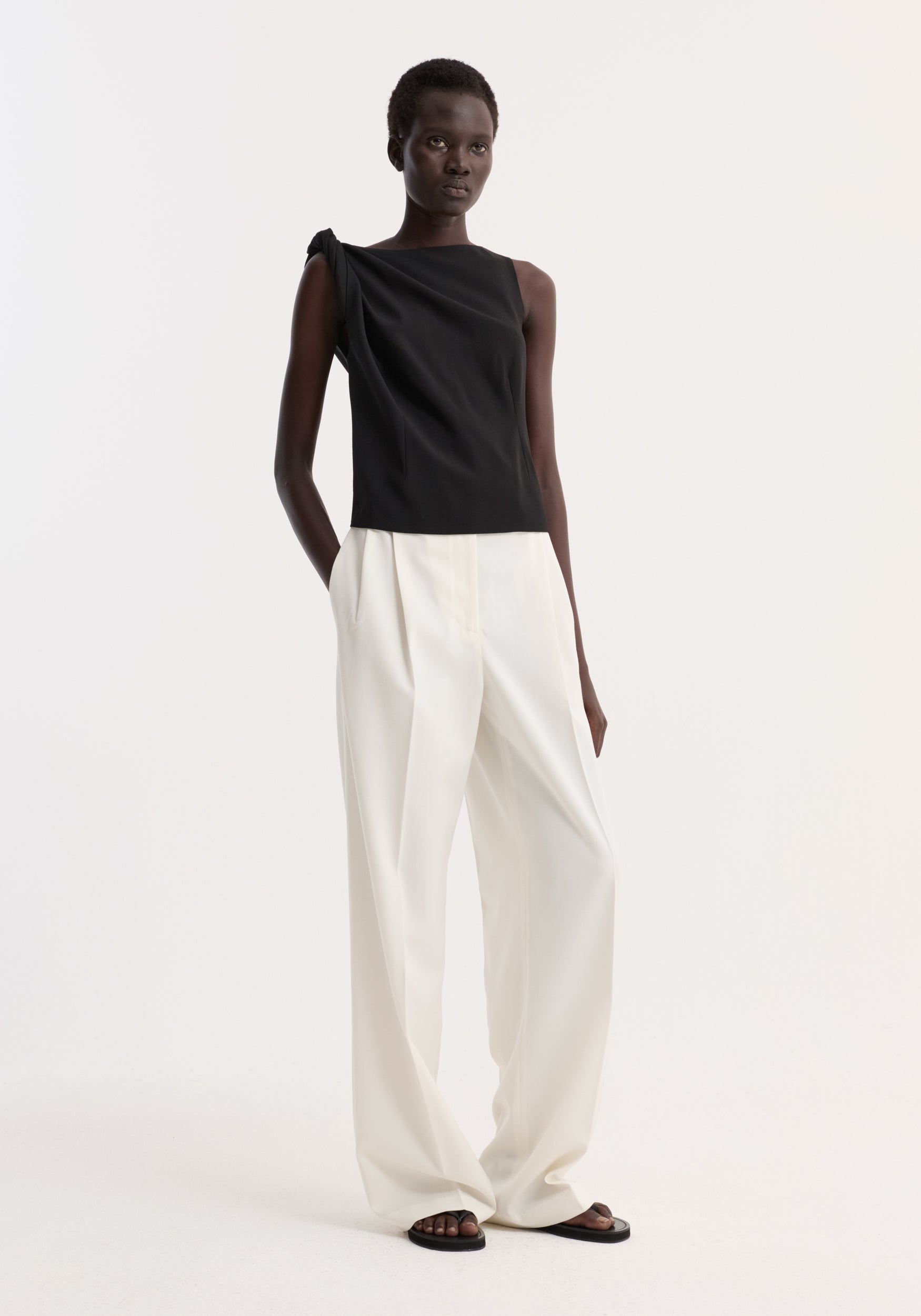 Model wearing a Draped linen silk top | black, featuring a twist detail at the shoulder, boat neck, and pleated side, paired with white trousers.