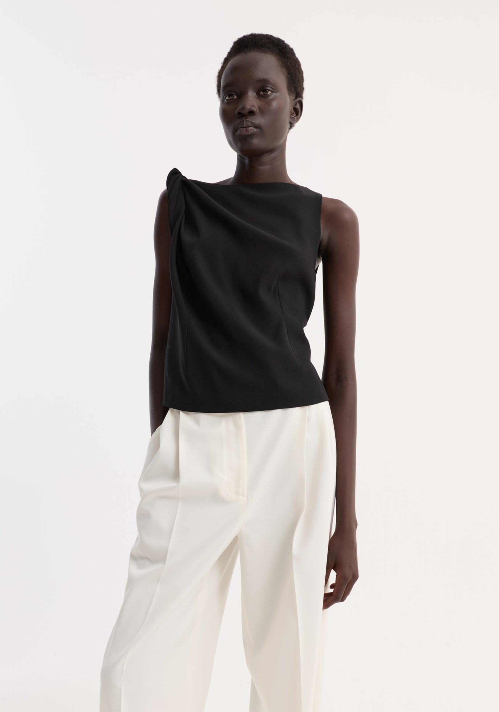 Model wearing the Draped linen silk top | black, featuring asymmetrical drape, twist detail on shoulder, boat neck, paired with wide-leg trousers.