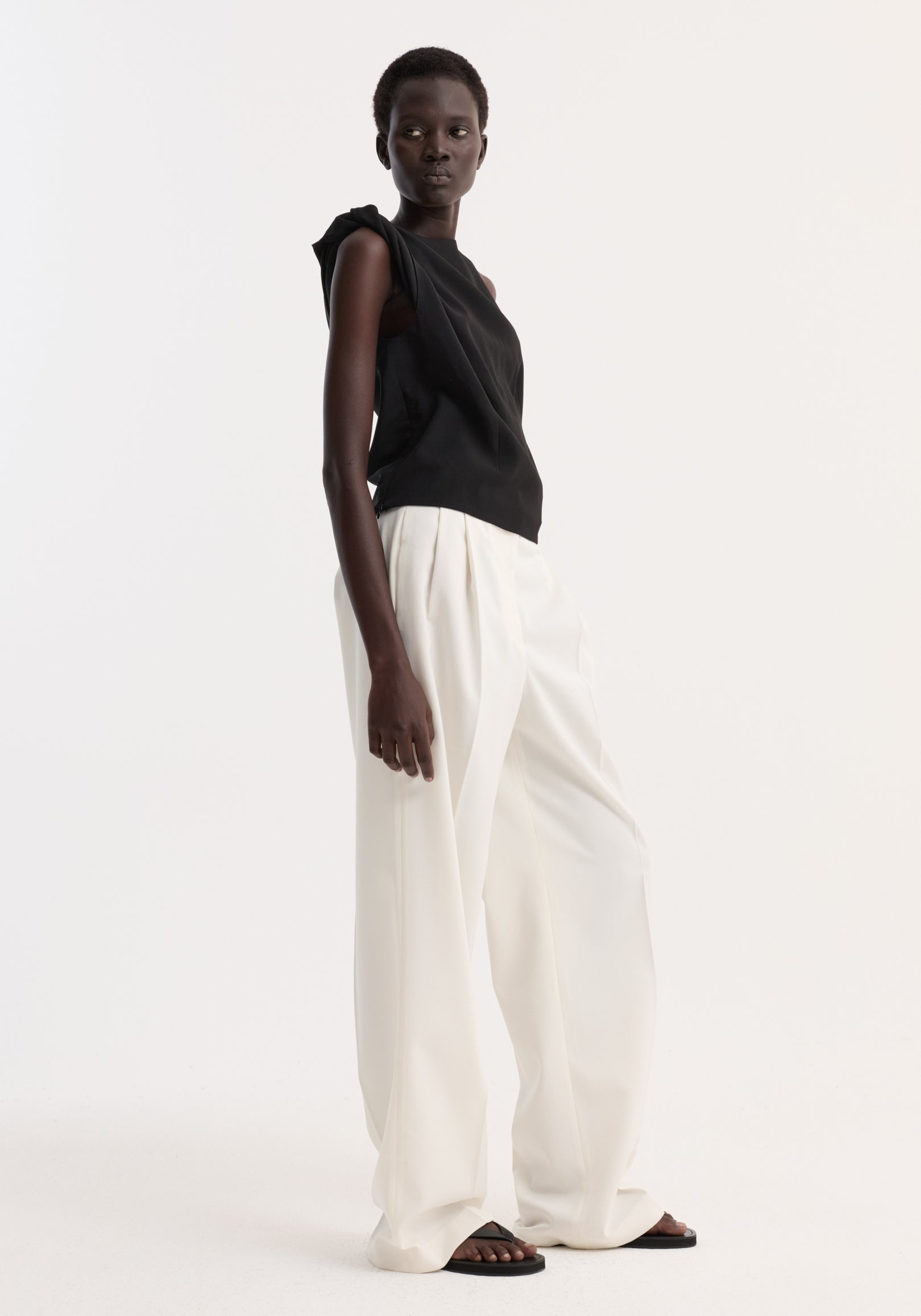 Model wearing Draped linen silk top | black with twist detail on one shoulder, boat neck, and pleated gathering. Paired with high-waisted white trousers.