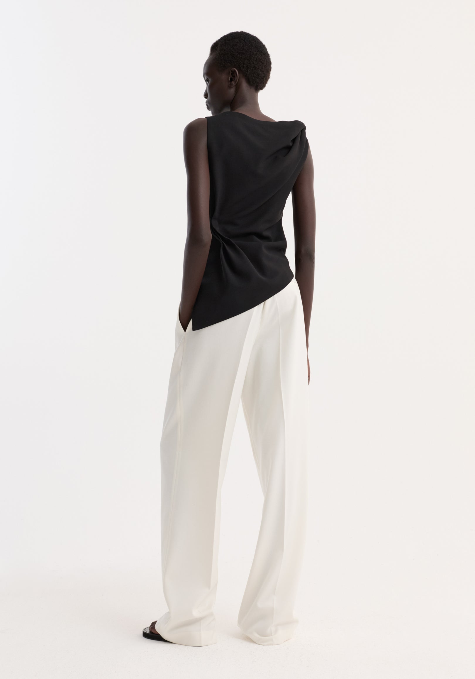 Draped linen silk top in black with a twist detail and boat neck, worn by a model with wide-leg trousers, showcasing an asymmetrical silhouette.
