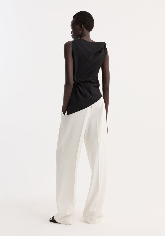 A person models the Draped linen silk top | black, showcasing its asymmetric drape, twist detail, boat neck, and gathered waist, paired with wide-leg trousers.