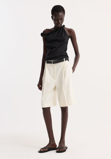 Person wearing a Sculptural cotton silk top | black, with a one-shoulder, twist detail, paired with high-waisted shorts and black flip-flops, standing relaxed.