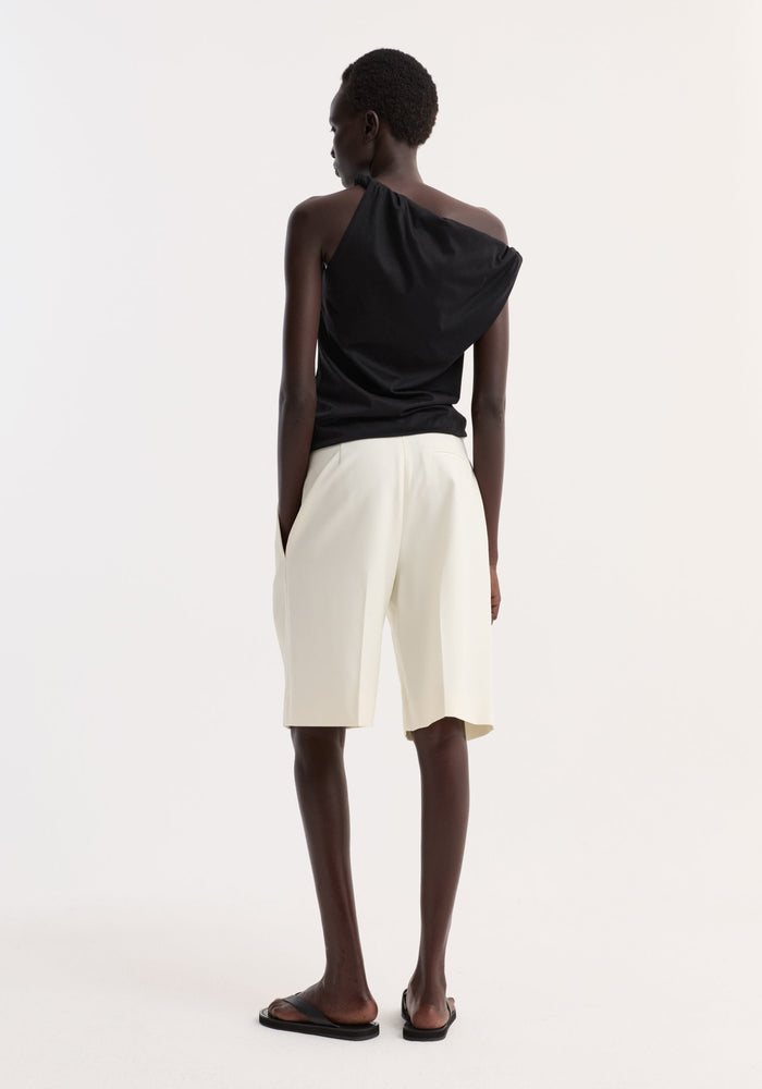Model in Sculptural cotton silk top with twisted strap and asymmetrical design, paired with high-waisted bermuda shorts. Hands in pockets, wearing flat sandals.
