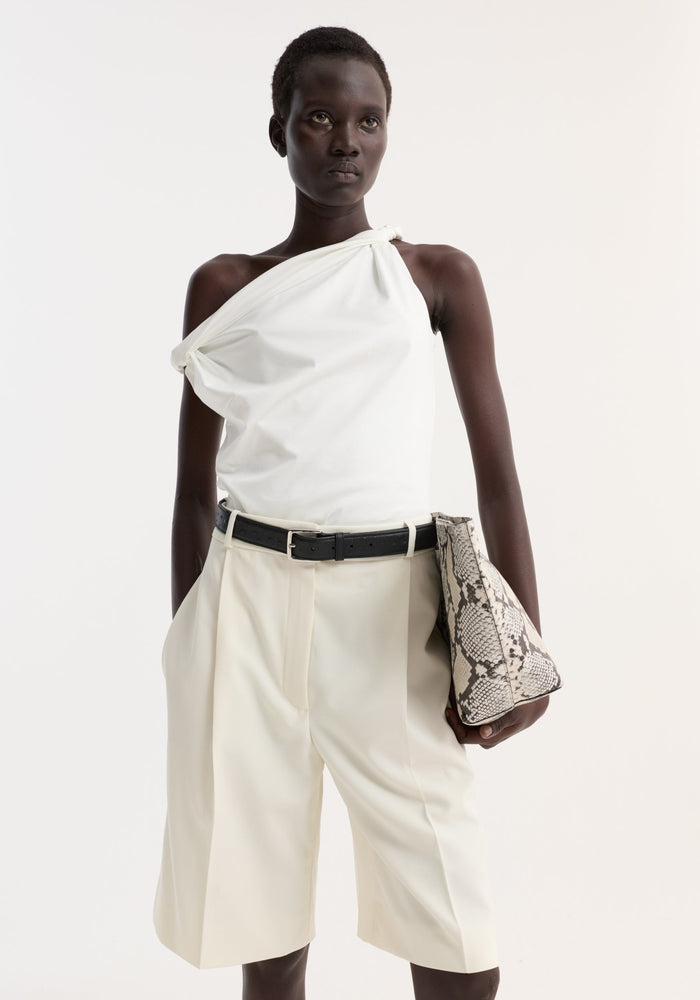 Woman wearing Sculptural cotton silk top with twisted strap, one-shoulder design, paired with high-waisted Bermuda shorts, holding a snakeskin-print clutch.